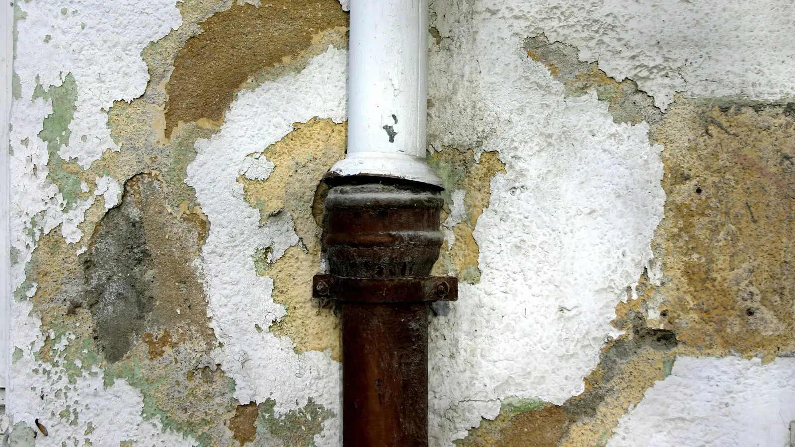 A rusted pipe on a wall with peeling paint and water staining around it